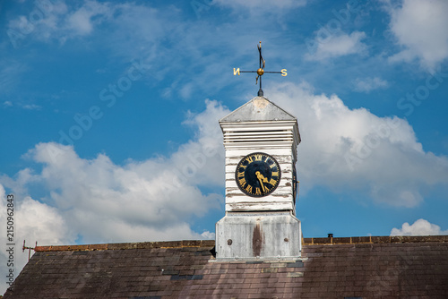 Weathered traditional clock tower with Roman numerals and weathervane against blue cloudy sky in rural England.