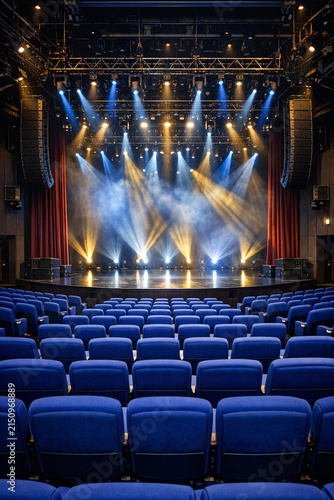 A modern theater with blue seats and stage lights set for a performance, empty of audience.