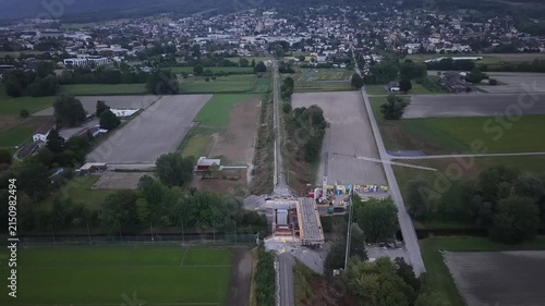 Forward aerial drone flight following a railway line across a river construction site where a new bridge is being built, with the railway continuing toward a town in the distance.