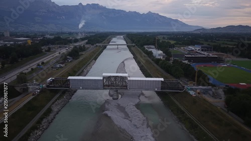 Forward drone flight toward a railway bridge undergoing renovation over the Rhine River, surrounded by extensive scaffolding and a temporary protective roof for construction work.