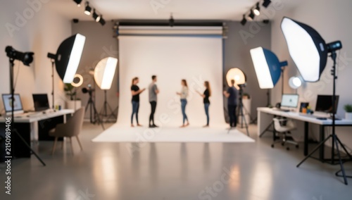 Out of focus wide shot of a professional film and photography studio interior with white cyclorama backdrop, softbox lighting kits, and production crew on set.