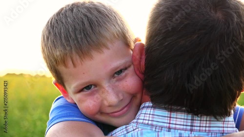 Young dad holding in hands his happy small son at countryside. Little smiling boy looking into camera and hugging his father against bright sunlight at background. Concept of family resting. Close up