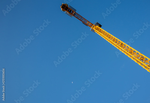 Yellow tower crane jib angled skyward against clear blue sky with moon visible