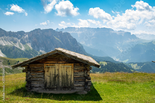 Konstfotografi Rustic wooden mountain hut on Seceda alpine meadow overlooking Ortisei in Dolomites, Italy, with dramatic mountain peaks and valley in background under bright blue sky with clouds