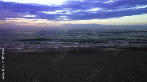Serene seascape at sunrise with gentle waves lapping on a dark, wet sand beach. The tranquil water reflects a dramatic cloudy sky