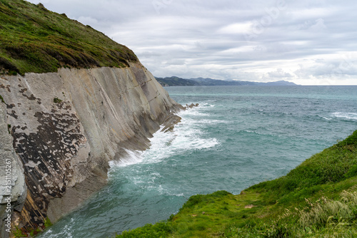 Dramatic geological formations of layered rock meet the churning turquoise waters of the Cantabrian Sea under a cloudy sky