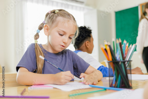 Schoolgirl writing with pencil in classroom, focused on learning and education during a school lesson with classmates, representing studying, homework, creativity, and childhood development.