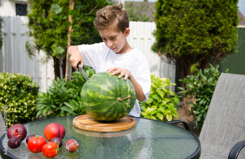 Cuadro en lienzo Young boy carefully cutting a whole watermelon on a wooden board outdoors