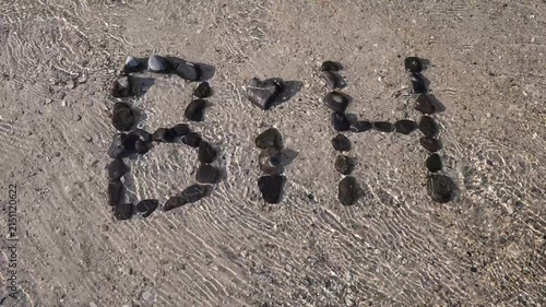 Stones arranged to spell BIH on a river bank splashed by water. Pebbles spelling the abbreviation for Bosnia and Herzegovina on the shore of a flowing river.
