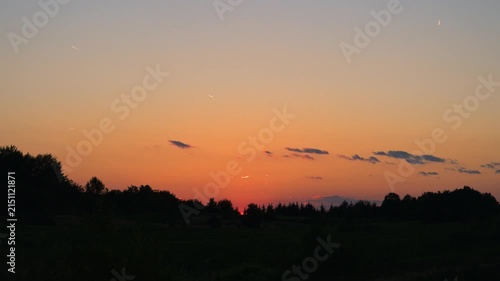 Vibrant sunset or sunrise with a warm glowing orange and red sky above a dark treeline silhouette of trees on a mountain. Colorful twilight sky with dramatic orange and red tones.