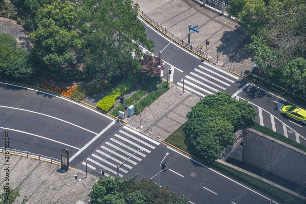 Obraz premium Aerial view of pedestrian crossings beside a landscaped road underpass ramp, flowering beds edging the descent into the underpass, a study in street geometry and lane use.