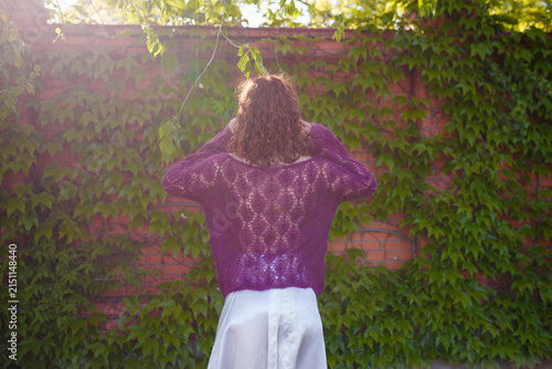Redhead girl in knitted purple transparent sweater posing at green ivy wall. Back view. Clothing and fashion concept