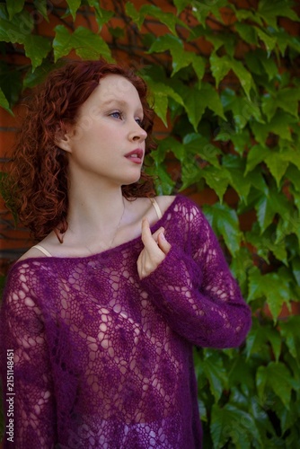 Redhead teenage girl in knitted purple transparent sweater posing at green ivy wall. Close up portrait. Clothing and fashion concept