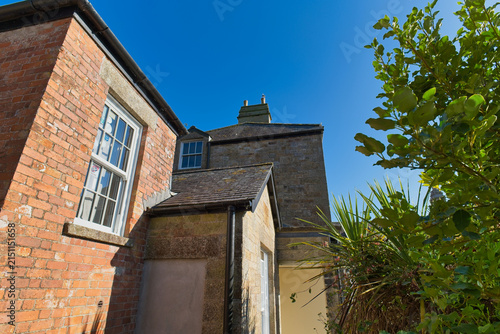 An old brick and stone building stands under a bright blue sky, with its detailed windows visible - Kenegie Manor, Cornwall, UK