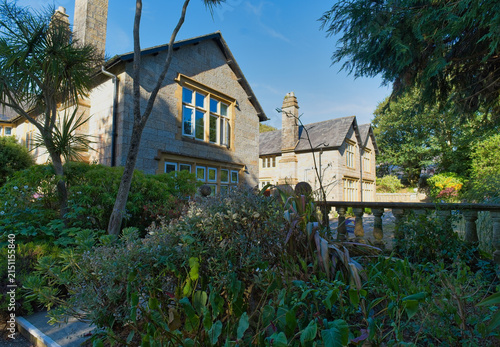 A charming stone building surrounded by lush greenery under a clear blue sky. The traditional architecture features prominent windows, with another building visible behind - Kenegie Manor, UK