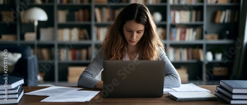 young woman working on an online research project at home, laptop screen facing away from camera, printed papers notebooks and books on desk, modern library interior, cinematic natural lighting,