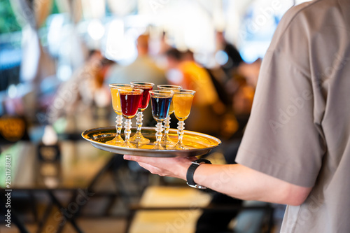 Принти на полотні A waiter carries a silver tray filled with colorful cocktail shots in elegant glasses at an outdoor restaurant