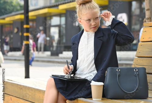 A little girl dressed as a businesswoman sits on a wooden bench outdoors