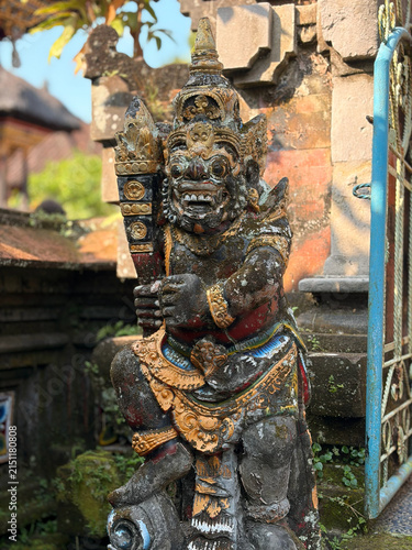 A stone statue of a fierce guardian figure, adorned with intricate carvings and colorful embellishments. The statue is set against a backdrop of lush greenery and traditional architecture.
