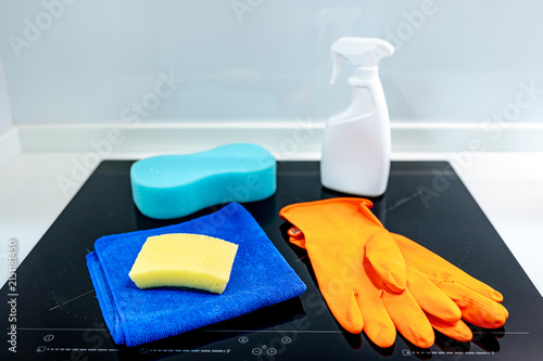 Cleaning tool products on countertop for home hygiene maintenance. Orange rubber gloves, blue microfiber cloth, sponge and detergent spray bottle on induction cooktop in modern kitchen