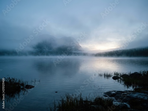 Mysterious Shape Emerges From Dense Mist Over A Calm Lake At Dawn, Creating An Ethereal And Serene Landscape.