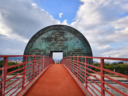 Symmetrical perspective view of the red pedestrian bridge passing through the giant ancient Chinese copper coin sculpture in Huize. HUIZE, YUNNAN, CHINA - JULY 30, 2026.