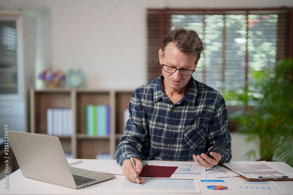 © Tj - Senior man analyzing financial reports and calculating household budget, managing personal finance with paperwork, smartphone, and laptop in a home office setting