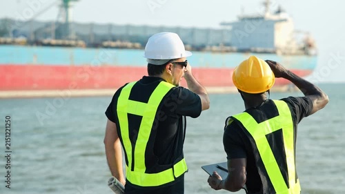 Professional maritime engineer team inspecting digital tablet with cargo ship background during port logistics safety operation at industrial shipping harbor coast