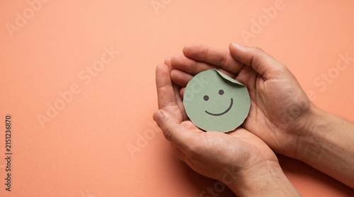 Hands holding paper smiley face on peach background symbolizing happiness positive emotion and well being care and support.