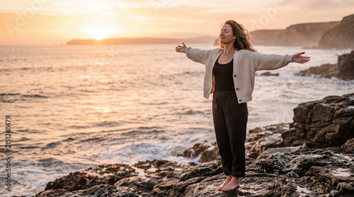A happy woman with outstretched arms embracing the sunset on a rocky ocean coastline.