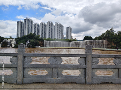 Scenic view of Kunming Waterfall Park and high-rise residential buildings seen through a traditional carved stone railing. KUNMING, YUNNAN, CHINA - JULY 31, 2026.
