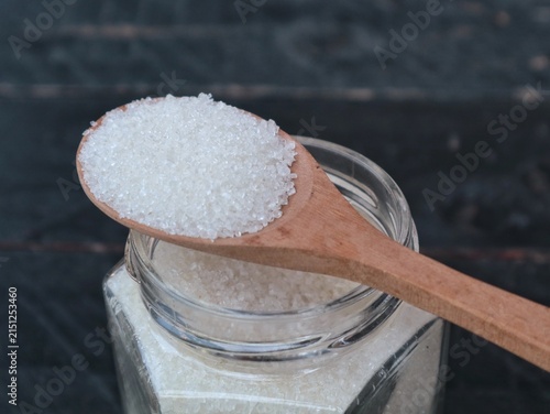 Fotografía White refined sugar granules in a clear glass jar with a wooden spoon placed on a dark rustic wooden background