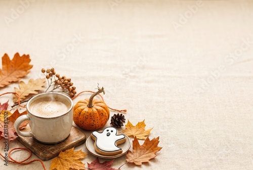 A cup of coffee sits next to a ghost-shaped cookie on a plate. A small pumpkin lies nearby on a table covered with autumn leaves. The setting captures a moment in fall
