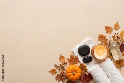 Different spa items are arranged neatly on a beige background. There are candles, towels, smooth stones, and autumn leaves. A small pumpkin adds to the seasonal decor