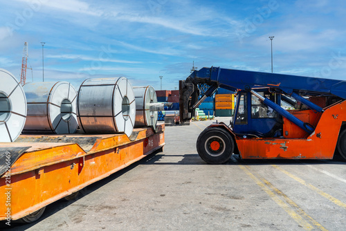 Reach stacker loading large steel coils onto a trailer at a container yard under a bright blue sky. Industrial logistics cargo handling, heavy machinery, and global shipping concept with strong detail