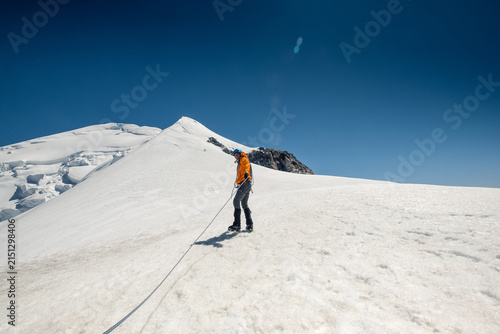 Climber In Orange Jacket Traversing Snowy Slope With Rope Mont Blanc Summit In Background Near Vallot Hut High Altitude Alpine Expedition In French Alps Outdoor Adventure Extreme Sports Trekking Hike