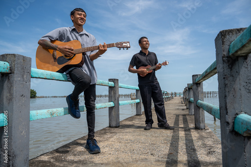 Two friends leaning on bridge railing playing acoustic guitar and ukulele. Two musicians enjoying playing music together on a coastal bridge.