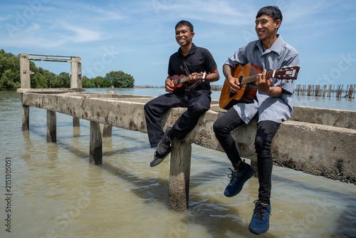 Two friends leaning on bridge railing playing acoustic guitar and ukulele. Two musicians enjoying playing music together on a coastal bridge.