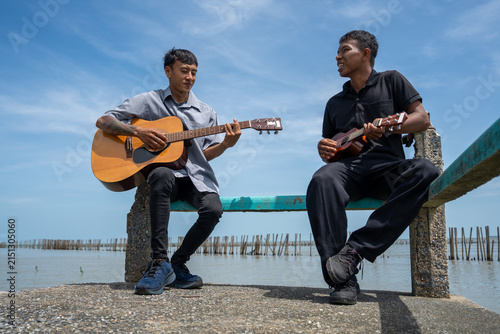 Two friends leaning on bridge railing playing acoustic guitar and ukulele. Two musicians enjoying playing music together on a coastal bridge.