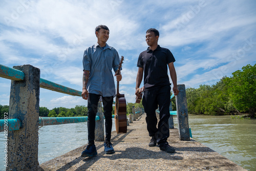 Asian friends carrying guitar and ukulele walking on coastal bridge. Two musicians strolling along a bridge near a mangrove forest while holding their instruments on a sunny day.