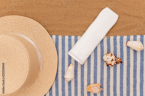 A straw hat, a white tube of cream on a blue and white striped blanket on the sand.