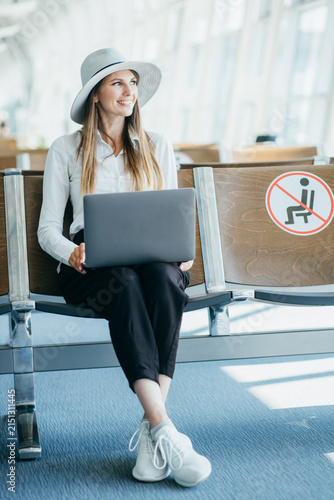 Woman wearing hat and white shirt works on laptop while waiting for flight in airport terminal