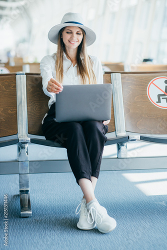 Young woman wearing a hat and white shirt works on her laptop while sitting in an airport waiting area
