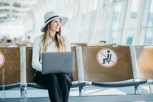 Woman wearing hat and white shirt works on laptop while sitting in airport waiting area with social distancing signs on seats