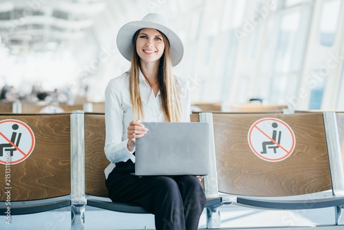 Smiling woman in hat holding laptop while sitting in airport waiting area with social distancing signs on seats