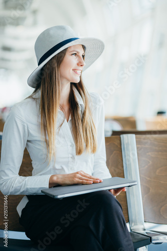Woman wearing hat and white shirt sits with laptop in airport terminal waiting area
