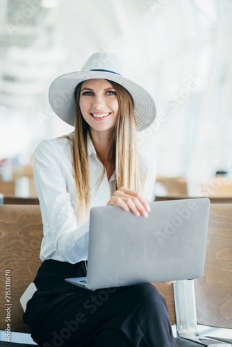 Young woman wearing a hat and white shirt sits with a laptop in an airport waiting area, looking at the camera with a smile
