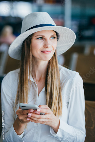 Woman wearing white shirt and hat holds smartphone in hands while looking away with a smile