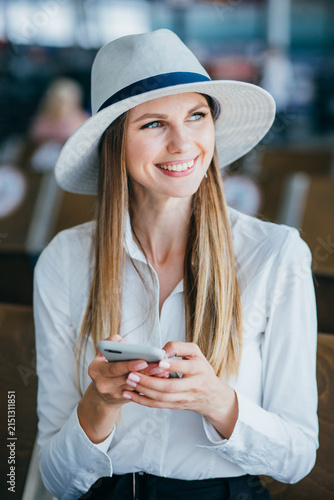 Young woman wearing fedora hat and white shirt, smiling while holding smartphone in hands, blurred background suggests airport or waiting area