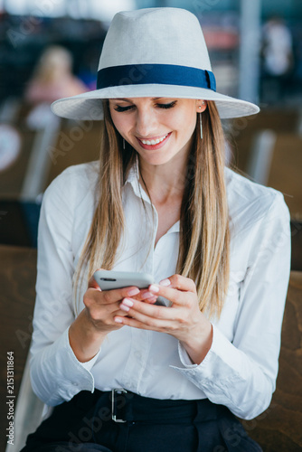 Young woman wearing a hat and white shirt smiles while looking at her smartphone, possibly waiting for a flight